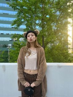 a young woman wearing a hat and cardigan standing next to a wall with trees in the background