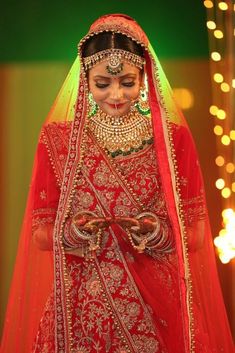 a woman in a red and gold bridal outfit with jewelry on her face, standing next to a string of lights