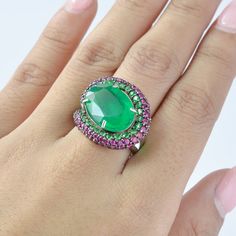 This exquisite ring features a stunning emerald center stone, surrounded by a radiant array of tsavorite and rhodolite garnet accent stones. The vibrant green emerald is elegantly framed by two concentric halos of glittering tsavorite and rich ruby, creating a captivating contrast that draws the eye. Set in a high-quality sterling silver band, this ring boasts a unique design that blends modern elegance with timeless sophistication. Perfect for special occasions or as a statement piece, this rin Fine Jewelry Green Emerald Ring With Accent Stones, Luxury Green Gemstones With Halo Setting, Luxury Green Sapphire Ring With Accent Stones, Oval Green Sapphire Ring With Halo Setting, Luxury Green Oval Gemstones, Luxury Green Rings With Gemstone Accents, Green Sapphire Ring With Halo Setting For Formal Events, Green Sapphire Ring With Halo Setting For Formal Occasions, Luxury Multi-stone Emerald Ring For May Birthstone