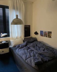 an unmade bed sitting next to a window in a room with white walls and blue carpet