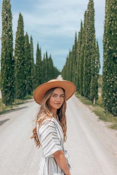 a woman wearing a hat standing on the side of a road with trees in the background