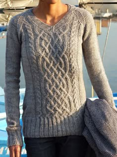 a woman standing in front of a boat wearing a gray sweater and blue jeans with her hand on her hip