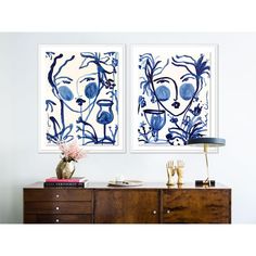 two blue and white paintings on the wall above a dresser with flowers in vases