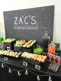 a black table topped with lots of cakes and cupcakes next to a chalkboard