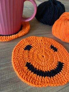 two crocheted pumpkin coasters next to a cup of yarn and a pink mug