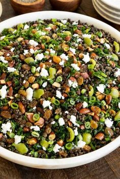 a white bowl filled with lots of food on top of a wooden table next to plates and utensils