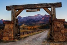 an old wooden gate in the middle of a dirt road with mountains in the background