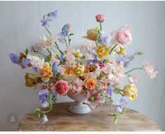 a vase filled with lots of different colored flowers on top of a wooden table next to a white wall