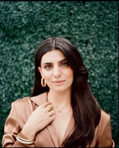 a woman with long black hair wearing gold hoop earrings and a tan jacket standing in front of a green wall