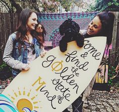 two girls holding a surfboard with writing on it and a dog in the middle