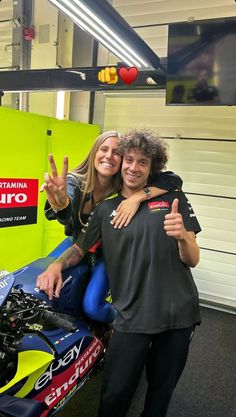 a man and woman standing next to a motorcycle giving the peace sign with their hands