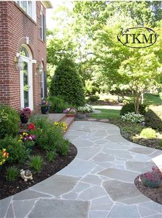 a stone walkway leading to a brick building with flowers and trees in the front yard