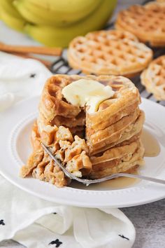 a stack of waffles on a plate with butter and bananas in the background