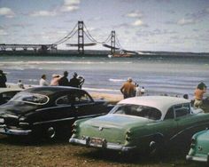 several old cars parked on the beach with people standing near them and a bridge in the background