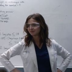 a woman wearing glasses stands in front of a whiteboard with writing on it and has her hands on her hips