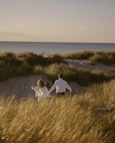 two people are walking through tall grass near the ocean and sand dunes, holding hands