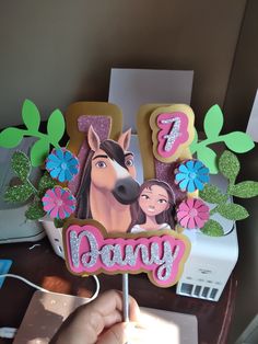 a hand holding a cake shaped like a horse with the name pony on it and flowers