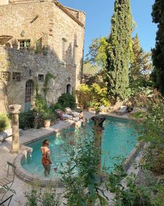 an outdoor swimming pool surrounded by trees and people in the water, with stone buildings behind it