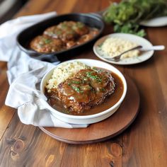 two plates with meat and gravy sitting on a wooden table next to other dishes