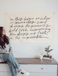 a woman sitting on top of a dresser in front of a wall hanging with writing