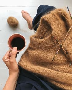 someone is holding a coffee cup with their feet up on the floor next to some knitting needles