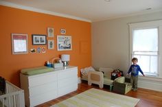 a young boy standing in the corner of a room next to a dresser and crib