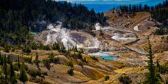 steam rises from the ground near trees and water in an area that is surrounded by mountains