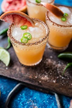 three glasses filled with drinks sitting on top of a wooden tray next to sliced fruit