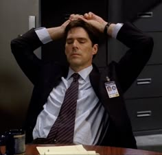 a man in a suit and tie sitting at a desk with his hands on his head