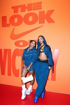 two women posing for the camera in front of an orange wall with the look of victory written on it