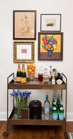 a table topped with bottles of wine next to pictures on the wall and vases filled with fruit