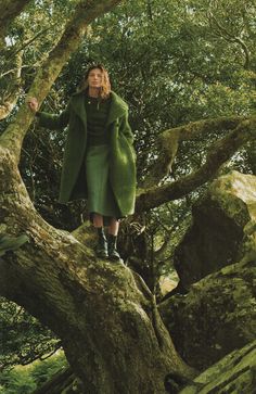 a woman standing on top of a tree in the middle of a forest wearing a green coat