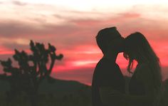 a man and woman kissing in front of a pink sky at sunset with cactus silhouettes