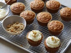 several cupcakes on a cooling rack with white frosting and toppings in bowls