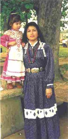 an old photo of a woman and child in native american clothing standing next to a tree