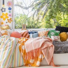 colorful pillows and blankets on a window sill in front of a sunny window with trees outside