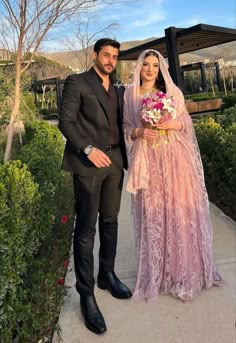 a man and woman standing next to each other in front of some bushes with flowers