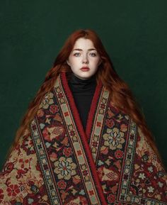 a woman with long red hair wearing a shawl and black turtle neck sweater, standing in front of a green background