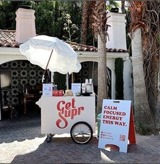 an ice cream cart with a white umbrella on the sidewalk next to palm trees and a sign that says get sup