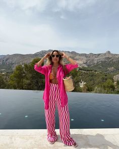 a woman in pink and white striped pants standing next to a body of water with mountains in the background