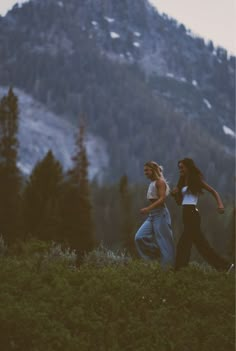 two women are walking in the grass near mountains