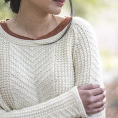 a woman with her arms crossed wearing a white sweater and brown leather collared necklace