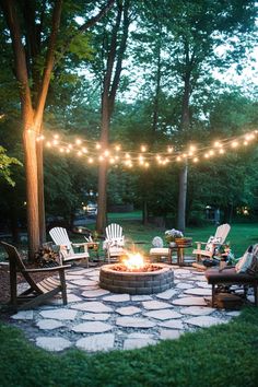 Backyard fire pit area surrounded by Adirondack chairs, lit by string lights. Front Yards Diy, Landscaping Around House, Cozy Backyard, Fire Pit Area, Backyard Lighting, Fire Pit Patio, Backyard Fire