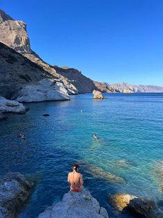 a woman sitting on top of a large rock next to the ocean with people swimming in it