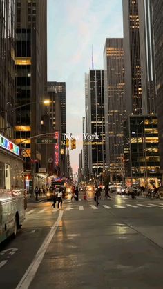 people are crossing the street in front of tall buildings at dusk, with lights on