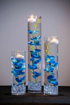 three tall glass vases with blue flowers and candles in them on a wooden table