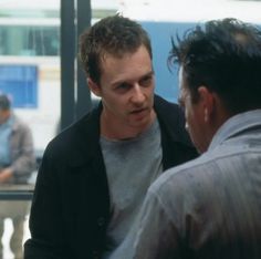 two men talking to each other in front of a bus station window with people walking by