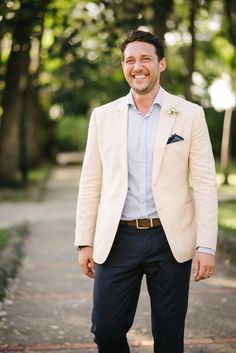 a man in a tan jacket and blue pants smiles at the camera while standing on a sidewalk