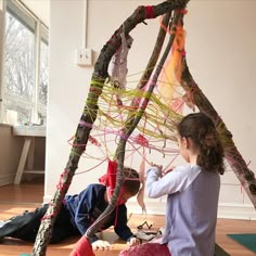 two children playing with yarn and sticks in front of an art project on the floor