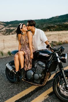 a man and woman kissing on the back of a black motorcycle with mountains in the background